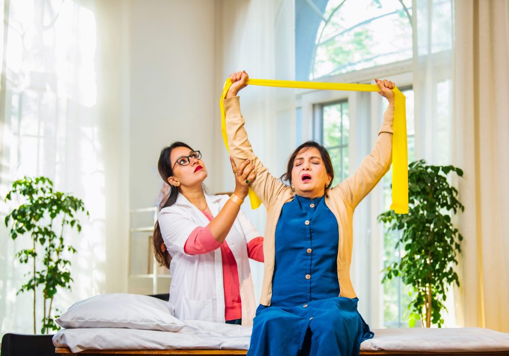 Senior Indian woman with shoulder pain learning resistance band exercise from female physiotherapist Indian Asian young female doctor, physiotherapist guiding senior woman patient with shoulder joint pain to perform resistance band, theraband exercise for strength, mobility rehabilitation in clinic