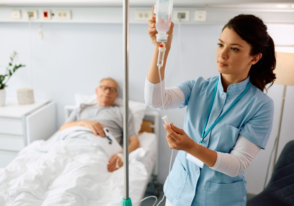 Female nurse taking care of hospitalized senior patient at intensive care unit. Nurse practitioner checking IV drip of elderly patient at hospital ward