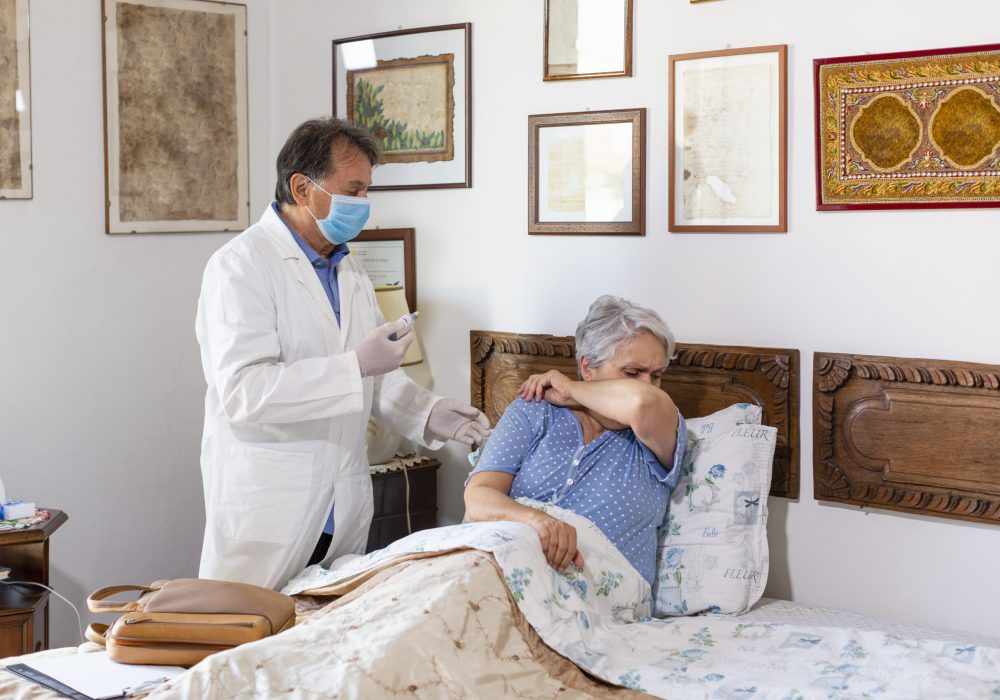 A doctor making a home visit to a senior woman patient. Woman coughing covering her mouth with her arm. Healthcare at home Senior woman receiving a vaccine from a visiting doctor.